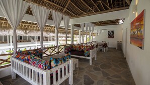 A covered outdoor seating area with white wooden benches and colorful cushions, a stone floor, and a thatched roof structure. - Ndoto House in Rafiki Tamu Dunia Resort (Watamu)