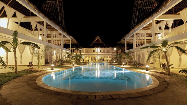 A nighttime view of a resort with a central pool, surrounded by palm trees and a thatched-roof building. - Ndoto House in Rafiki Tamu Dunia Resort (Watamu)