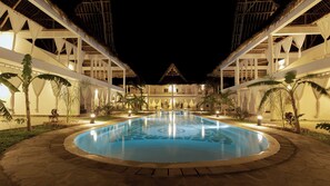 Een nachtzicht van een resort met een centraal gelegen zwembad, omgeven door palmbomen en een gebouw met rieten dak.