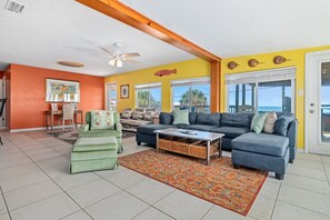 A spacious living room with a large sofa, a coffee table, and a chair. There are windows with a view of the ocean.