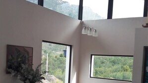 Una habitación con un sofá azul, una mesa de comedor con sillas, una lámpara y ventanas amplias con vistas a montañas.