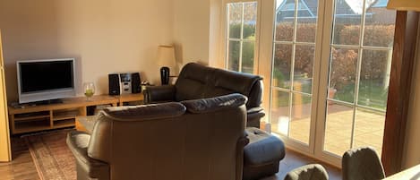 A living room with a leather sofa, a TV, speakers, and a lamp. There is a large window with a view of a garden and houses.