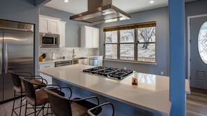 A modern kitchen with a central island, stainless steel appliances, and a large window with a view of trees. - Lakefront 6BR Retreat w Dock & Game Room (Burnet)