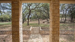 A stone-paved pathway leading to a covered area framed by stone pillars, surrounded by a natural landscape with trees and a grassy area. - Lakefront 6BR Retreat w Dock & Game Room (Burnet)