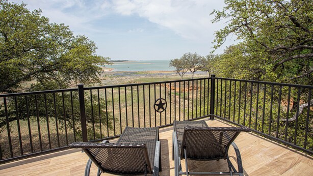 A balcony with two wicker chairs facing a beach view, a star emblem on the railing, and a wooden deck. - Lakefront 6BR Retreat w Dock & Game Room (Burnet)