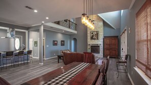 A spacious living area with a large wooden table, a fireplace, and a high ceiling with exposed beams. - Lakefront 6BR Retreat w Dock & Game Room (Burnet)
