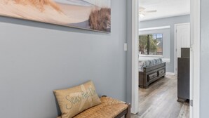 A hallway with a large beach-themed painting, a bench with a cushion, and a view into a bedroom with a bed and dresser.