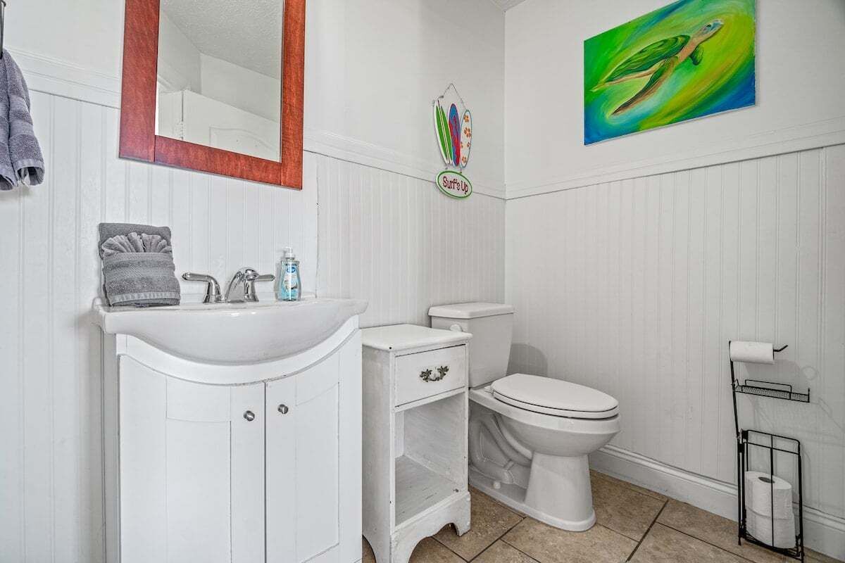 Une salle de bain avec un meuble-lavabo blanc, des toilettes, un lavabo et un miroir. Il y a un tableau au mur et un porte-serviettes.
