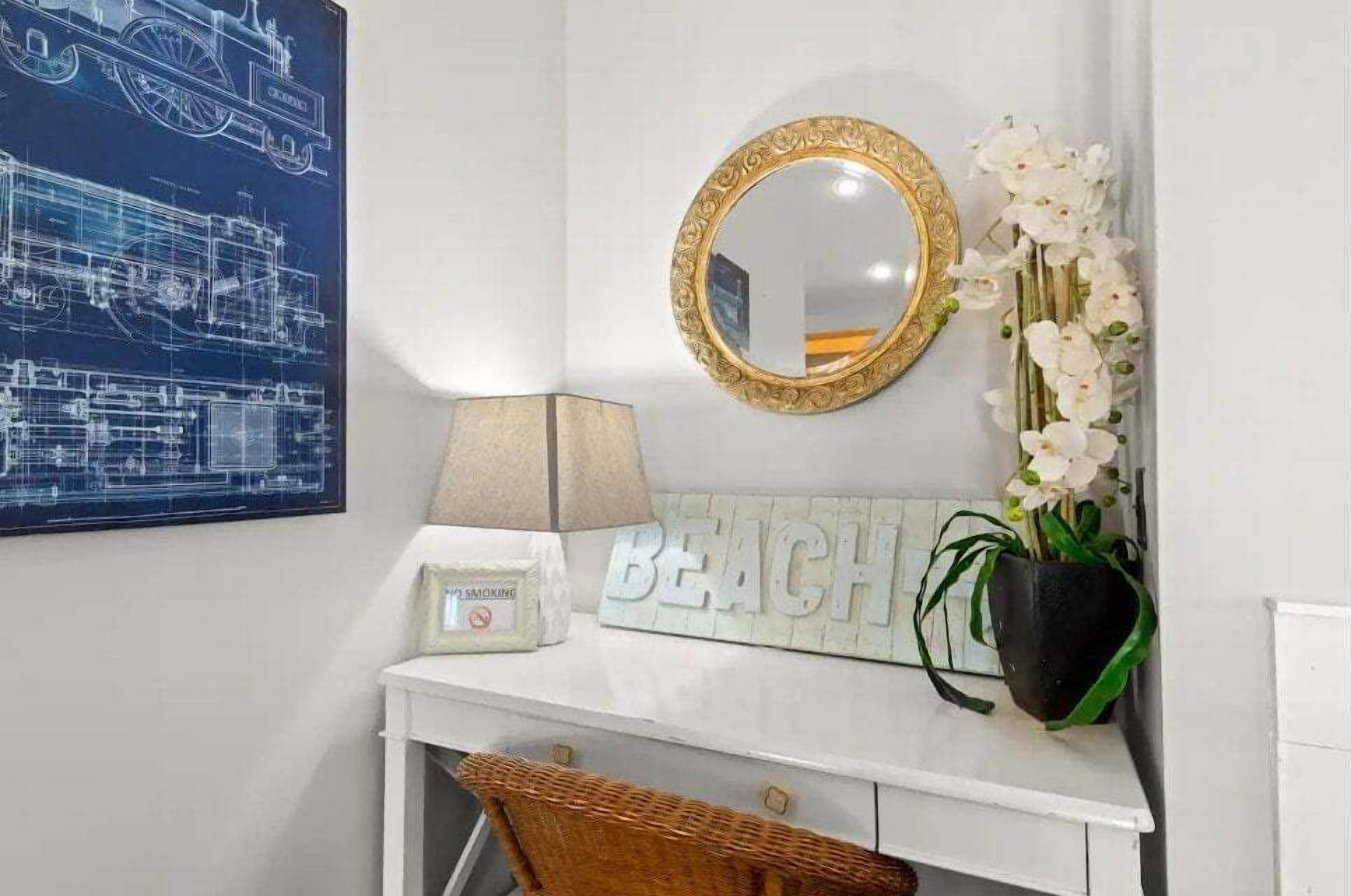 A bathroom with a white vanity, a framed blueprint, a 'BEACH' sign, a mirror with a gold frame, a lamp, and a potted plant.