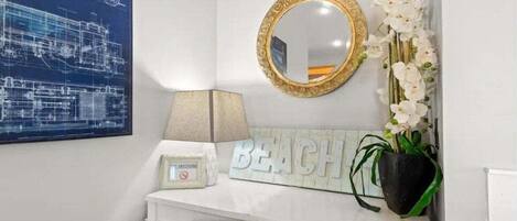 A bathroom with a white vanity, a framed blueprint, a 'BEACH' sign, a mirror with a gold frame, a lamp, and a potted plant.