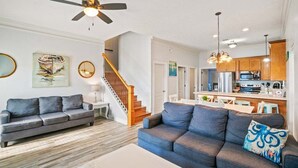 A modern living room with a gray sofa, a blue sofa, and a white coffee table. There is a staircase leading to a kitchen area with wooden cabinets and a dining table. - Fall Deals + Snowbird Rates | Coastal Stay Near the Beach (Panama City)