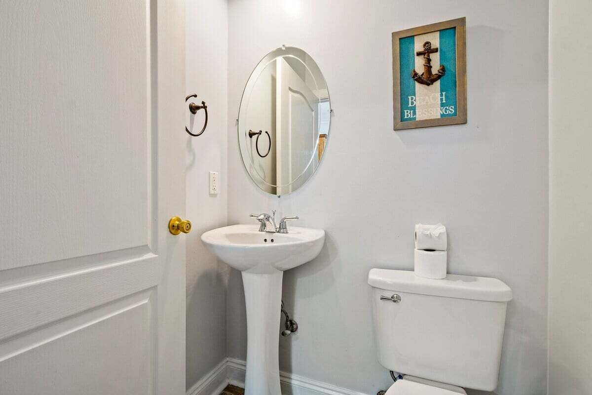 A bathroom with a pedestal sink, a toilet, a round mirror, and a framed picture on the wall.