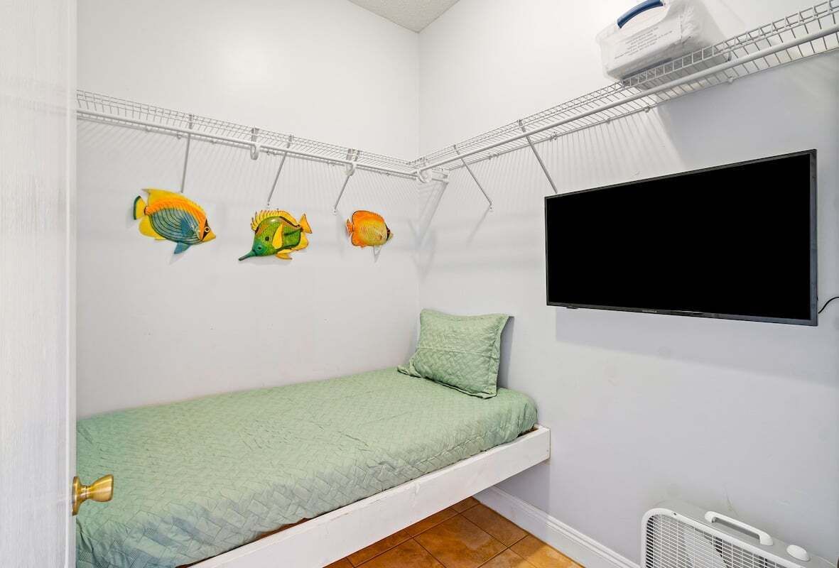 A small, single-bed room with a green bedspread, a mounted TV, and a wall-mounted drying rack with colorful fish decorations.