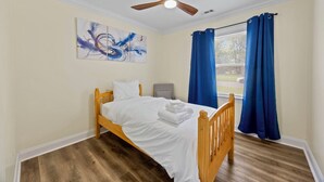 Une chambre à coucher avec un lit en bois, un ventilateur de plafond, une fenêtre avec des rideaux bleus et des œuvres d’art abstraites accrochées au mur.