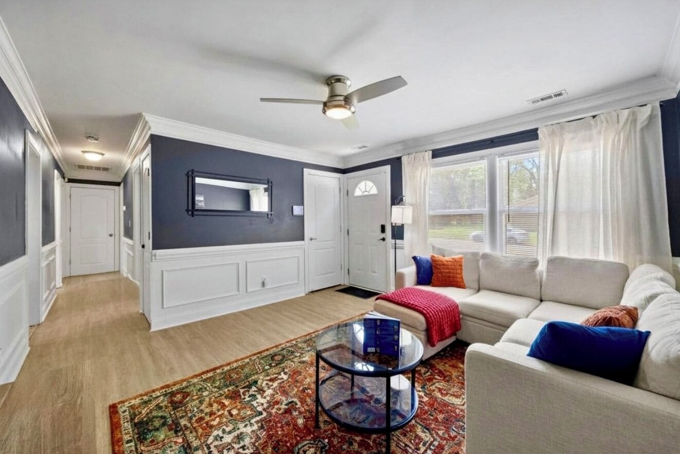 A living room with a sofa, a coffee table, a ceiling fan, and a large window with curtains.