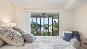 Une chambre avec un grand lit, une table de chevet, une lampe et une vue sur un balcon équipé d’une table et de chaises.