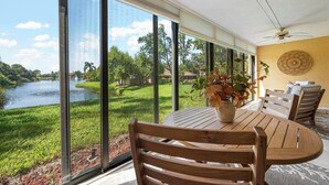 Une pièce dotée de grandes fenêtres donnant sur un lac et une végétation luxuriante, une table en bois avec des chaises et un ventilateur de plafond.