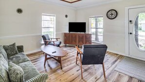 A living room with a sofa, two chairs, a coffee table, a TV on a wooden cabinet, and a door with a circular window.