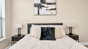 A bedroom with a bed, two bedside tables, a wall-mounted abstract painting, and a window with blinds.
