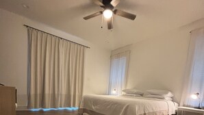 Un dormitorio con cama, mesita de noche, ventilador de techo y ventana con cortinas.