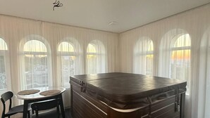 Una habitación con un jacuzzi de madera, una mesa de comedor con sillas y grandes ventanales con cortinas translúcidas.