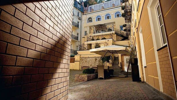 Un estrecho callejón con un muro de ladrillo a la izquierda y un edificio amarillo a la derecha. Hay una zona de estar al aire libre cubierta, con plantas y una escalera que conduce a los pisos superiores del edificio.