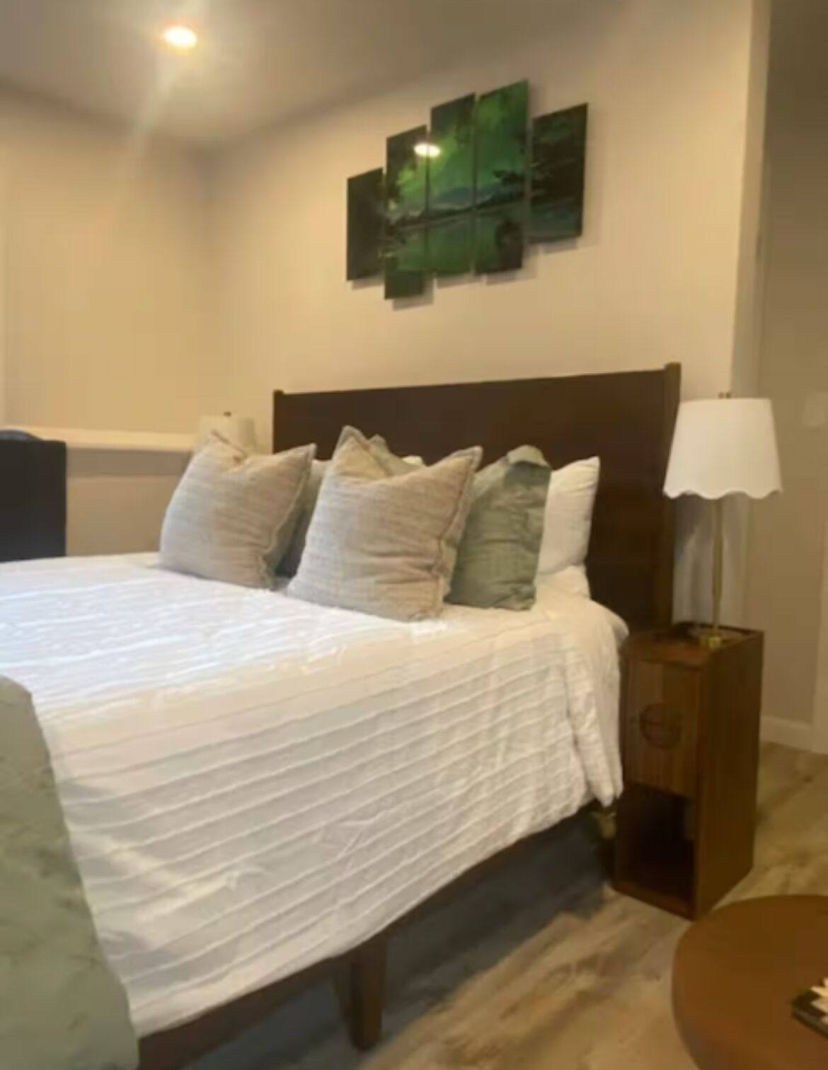 Una cama bien hecha con edredón blanco y varios almohadones, una mesita de noche de madera con una lámpara, y una obra de arte colgada en la pared sobre la cama.