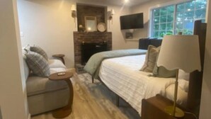 A cozy bedroom with a bed, a sofa, a small table, a lamp, a fireplace, and a TV. - Peaceful Pines Haven~ cozy studio near the ponds (Hanson)