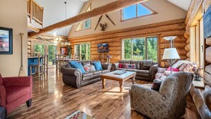 A spacious log cabin interior with a high ceiling, wooden beams, a large living area with leather sofas, a dining area, and large windows offering views of the outdoors.