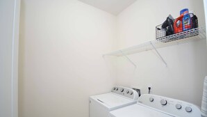 A laundry room with a front-loading washer and dryer, a metal shelf above the dryer holding cleaning supplies, and a white wall.