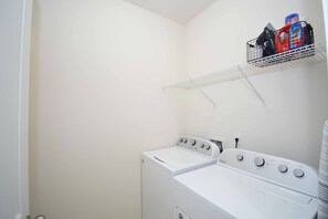 A laundry room with a front-loading washer and dryer, a metal shelf above the dryer holding cleaning supplies, and a white wall. - Getaway on Mclernon - Top Notch Mid Term Stay (Johns Island)