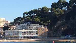 A coastal building with multiple floors, a beach in the foreground, and trees on a hillside in the background. - Rosamar Maxim Adults Only (Lloret de Mar)