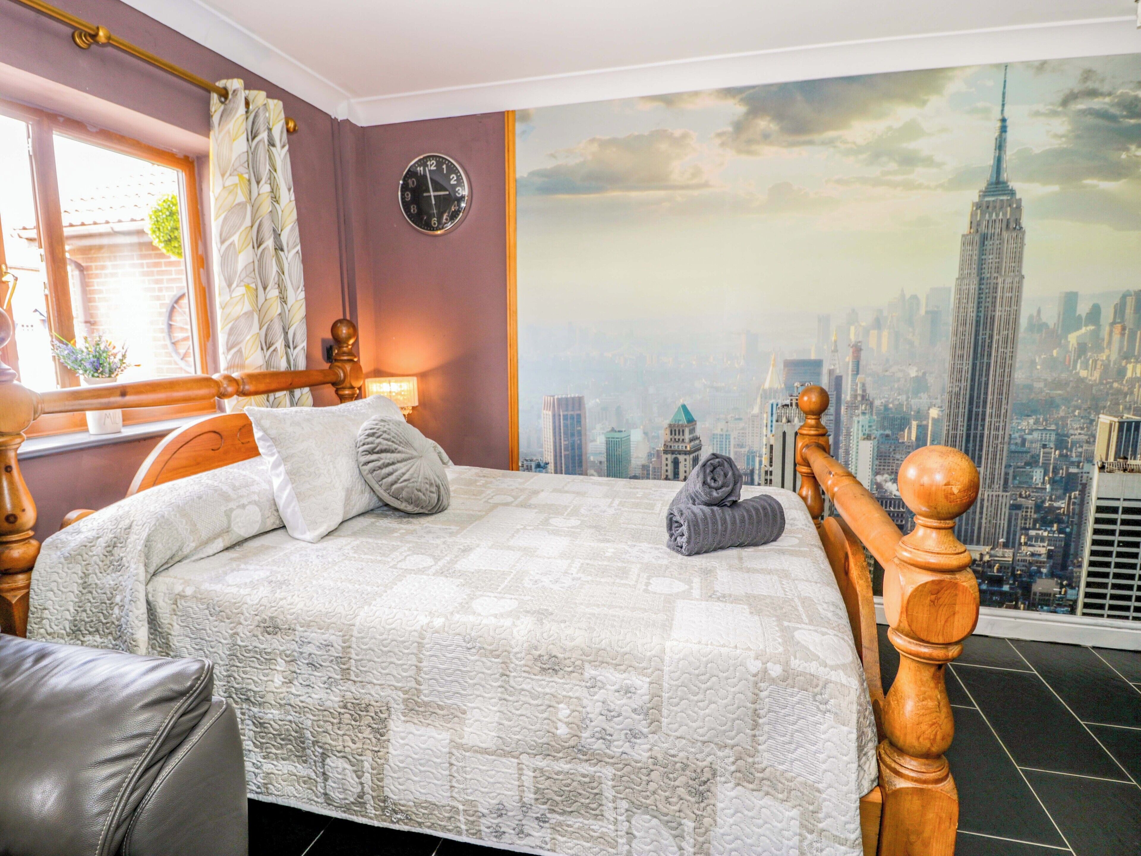 Ein Schlafzimmer mit einem Bett, einer Uhr, einem Fenster mit Vorhängen und einem Stadtbild-Wandgemälde.