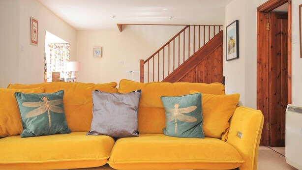 A yellow sofa with dragonfly pillows in a room with a wooden staircase and a radiator. - Rosewin Farmhouse (Wadebridge)