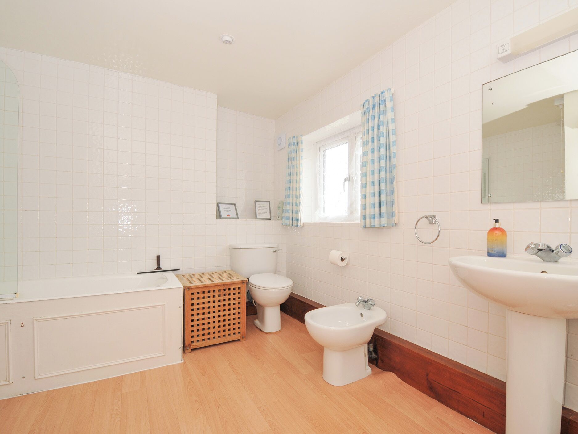 Un baño con paredes de azulejos blancos, suelo de madera, bañera, inodoro, bidé y lavabo.