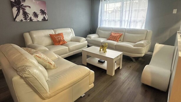 A living room with two white leather sofas, a white coffee table, and a white armchair. There is a painting on the wall and a window with curtains.