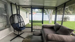 A covered patio with a grey sofa, a black mesh chair, and a small ottoman.