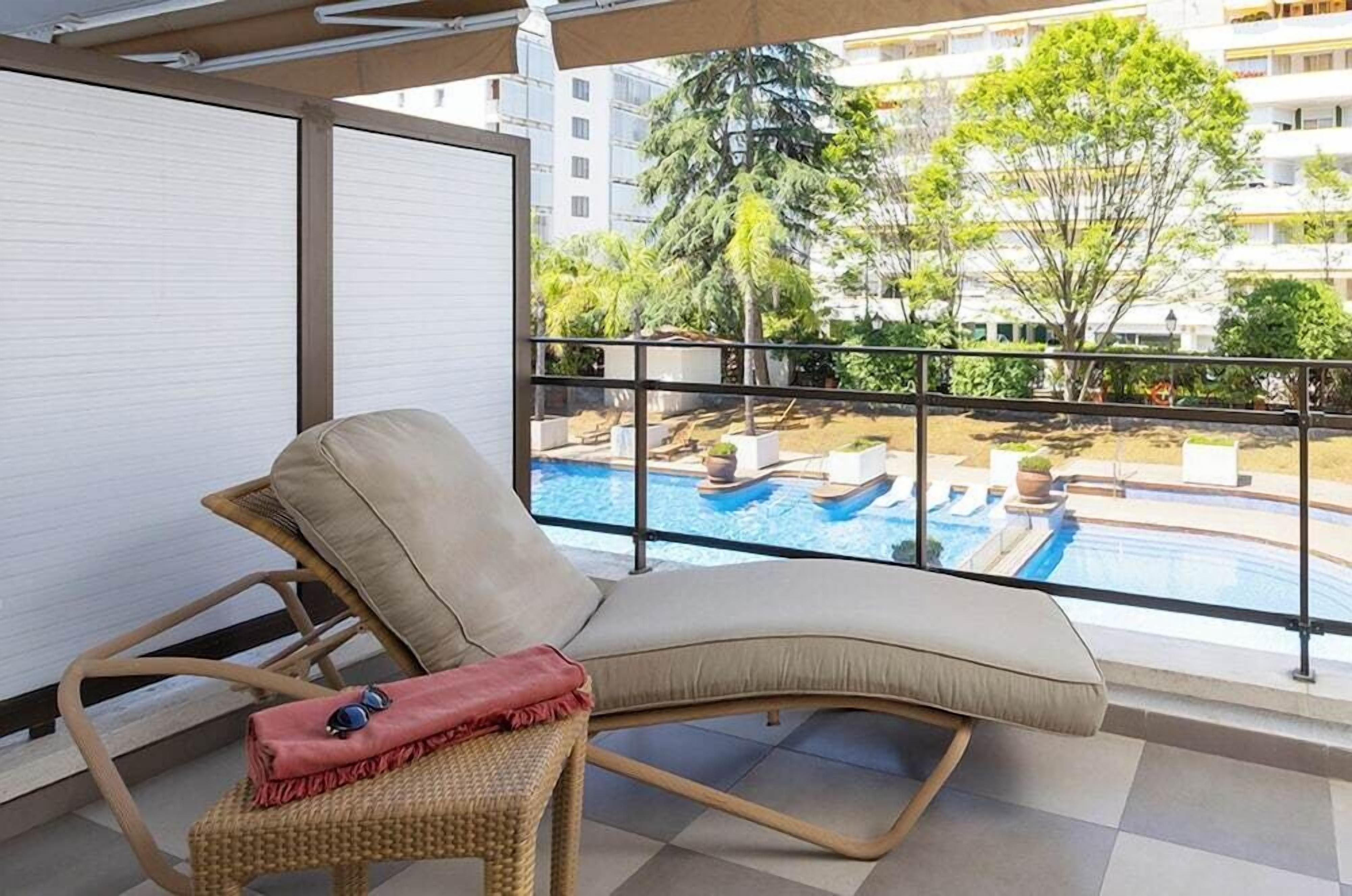A balcony with a lounge chair, a small table, and a view of a swimming pool and greenery.