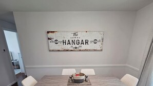 A dining area with a marble floor, a wooden table, and six white chairs. A rustic sign with the text "The HANGAR" is mounted on the wall. - Welcome to The Hangar, a newly remodeled travel-themed stay, 4 minutes from BDL. (Windsor Locks)