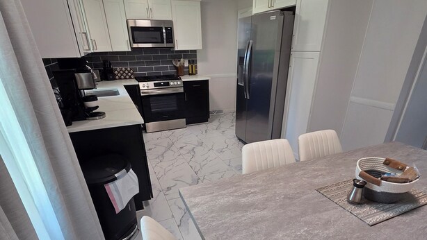 A modern kitchen with white cabinets, a stainless steel refrigerator, a microwave, and a stove. There is a dining table with chairs and a coffee maker on the table. - Welcome to The Hangar, a newly remodeled travel-themed stay, 4 minutes from BDL. (Windsor Locks)