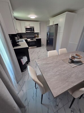 A modern kitchen with white cabinets, a stainless steel refrigerator, a microwave, and a stove. There is a dining table with chairs and a coffee maker on the table. - Welcome to The Hangar, a newly remodeled travel-themed stay, 4 minutes from BDL. (Windsor Locks)