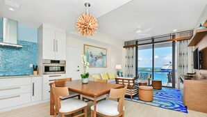Eine moderne Küche mit weißen Schränken, einem eingebauten Ofen und einer blauen Spritzschutzplatte. Ein Essbereich mit Holztisch und -stühlen lädt zum Verweilen ein, und ein großes Fenster bietet einen Blick auf den Ozean.
