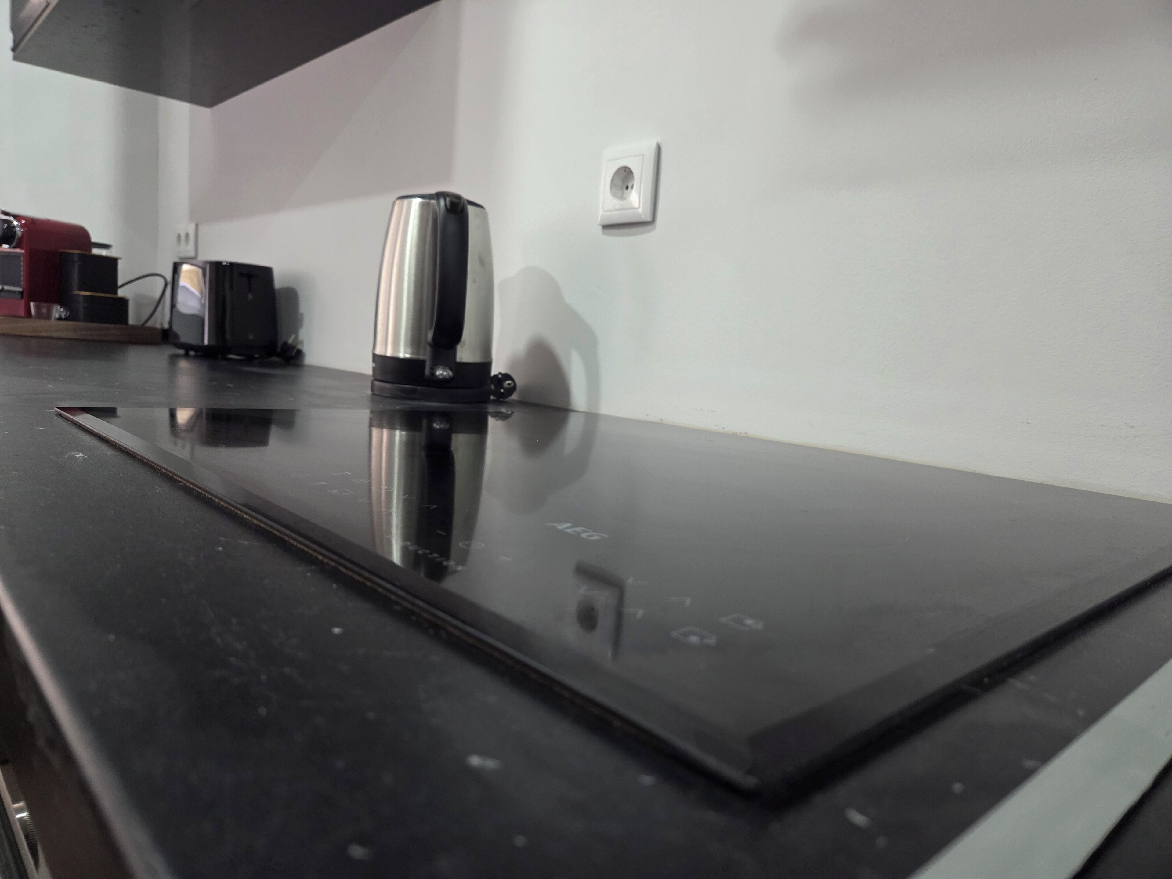 A kitchen counter with a stainless steel kettle, a coffee maker, and a power outlet on the wall.