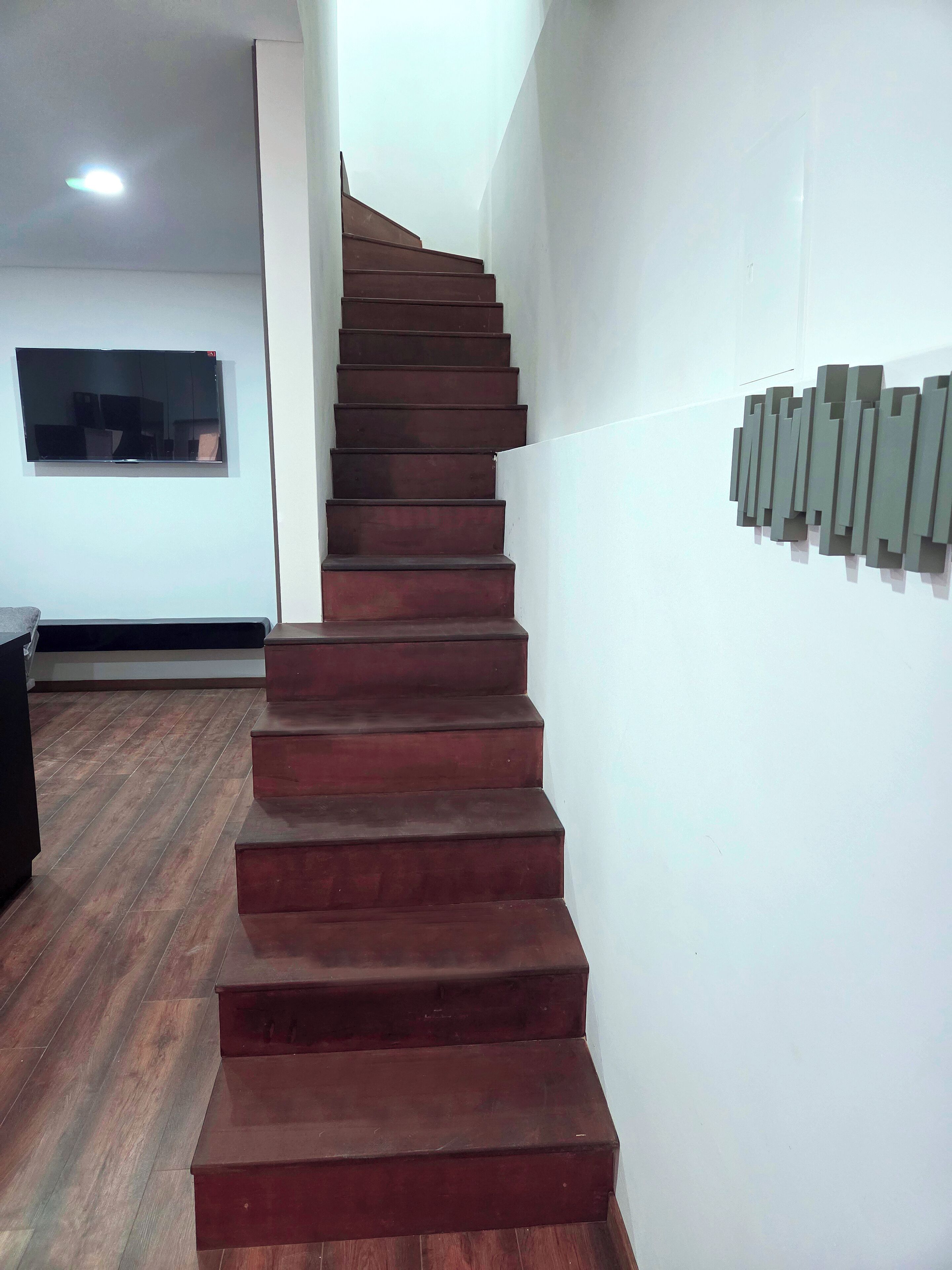 A staircase with wooden steps leading upwards, a flat-screen TV mounted on the wall, and a wall-mounted light fixture.