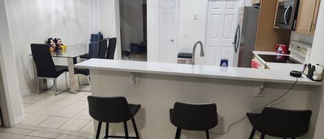 A modern kitchen with a central island, black bar stools, a dining area with chairs, and a visible microwave.
