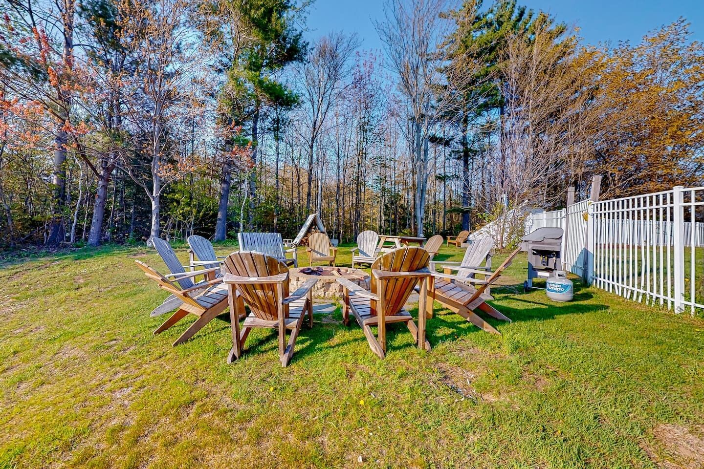 A backyard with a wooden table and chairs, surrounded by trees and a white fence.