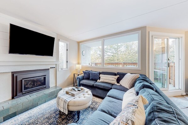 A modern living room with a fireplace, a large flat-screen TV, a sofa set, and a view through large windows. - Bridges 77 at Sugarbush (Warren)