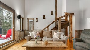 A cozy living room with a wooden staircase, a sofa set, a coffee table with a vase, and a large window overlooking a snowy deck. - Hidden Birch at Sugarbush (Fayston)
