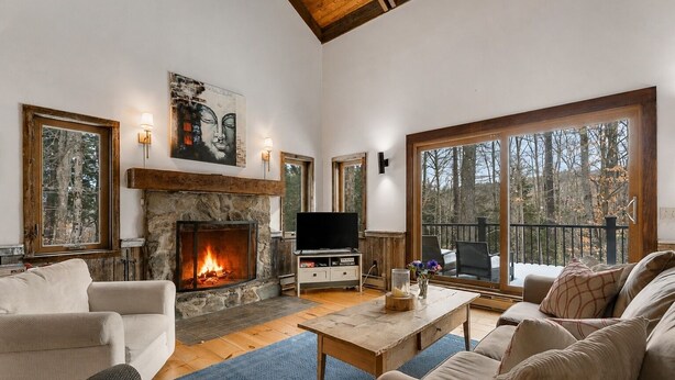 A cozy living room with a fireplace, a television, and a large window overlooking a forest. - Hidden Birch at Sugarbush (Fayston)