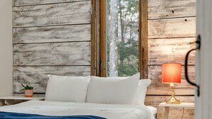 A bedroom with a wooden wall, a bed with white bedding, a blue throw blanket, a nightstand with a lamp, and a small potted plant. - Hidden Birch at Sugarbush (Fayston)